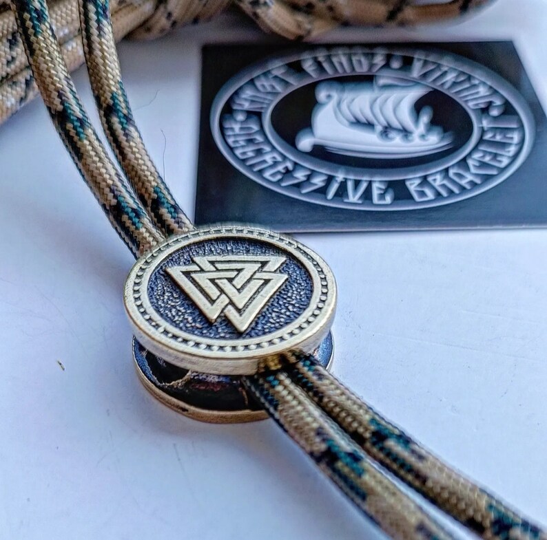 Op de afbeelding: Een bronzen paracord-schuif met een cirkelvormig ontwerp met een gestileerde driehoekige knoop. De schuif is bevestigd aan een paracord met een camouflagepatroon. De tekst "What Finds Vikings Aggressive Bracelet" is zichtbaar op een zwarte sticker op de achtergrond.