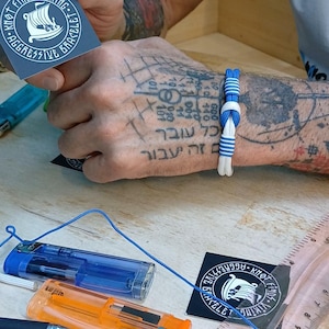 May include: A blue and white paracord bracelet featuring a central knot design. The bracelet is displayed alongside a clear plastic ruler showing measurements in inches and centimeters, two lighters, and a square card with a Viking ship logo.