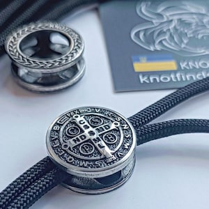 Puede incluir: Un amuleto de metal plateado con un diseño de medalla de San Benito. El amuleto está unido a un cordón negro.