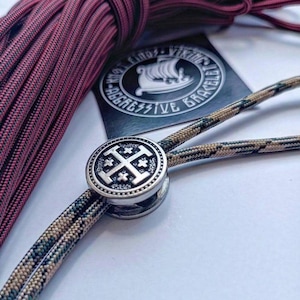 Puede incluir: Primer plano de una pulsera con un cierre plateado con un diseño de cruz. La pulsera está hecha de un cordón con estampado de camuflaje. Un haz de cordón burdeos y negro está en el fondo, junto con un logotipo en blanco y negro.