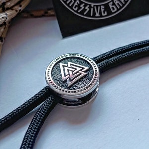 Pode incluir: Um bracelete de paracord preto com uma conta de metal prateada com um design em relevo de um nó celta, conhecido como Valknut.