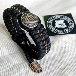 May include: Black paracord bracelet with a brown and gold metal charm featuring a Viking symbol. The charm is surrounded by a decorative knot. The bracelet is tied with a brown paracord. The bracelet is labeled "Knot Finds - Viking Aggressive Bracelet".