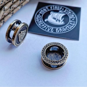 Op de afbeelding: Twee metalen kralen voor een paracord armband. E&eacute;n kraal heeft een gevlochten ontwerp en de tekst "Knot Finds". De andere kraal heeft een Viking symbool en de tekst "Each Knot Finds Viking Aggressive Bracelet".