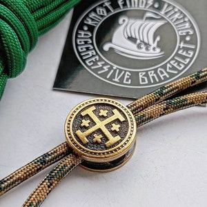 Op de afbeelding: Een gouden metalen kraal met een kruisontwerp. De kraal is bevestigd aan een bruin en groen paracord armband. Het armband is in een knoop geknoopt en heeft een label met de tekst "Aggressive Knot Finds Viking Brave Bracelet".