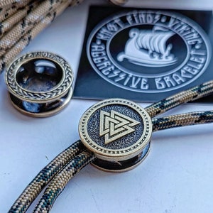 Op de afbeelding: Een close-up van een gouden metalen armband met een gevlochten koord. De armband heeft een ronde bedel met een Keltisch knoopontwerp. De bedel is bevestigd aan het koord met een metalen ring. De armband ligt op een wit oppervlak.
