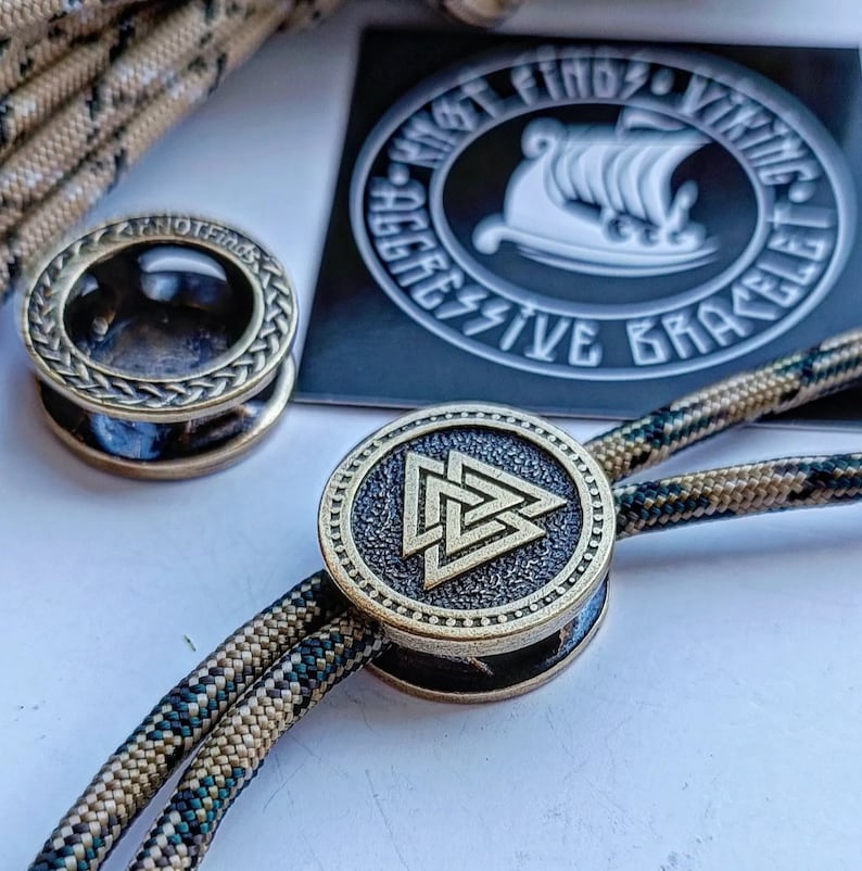 Op de afbeelding: Een close-up van een gouden metalen armband met een gevlochten koord. De armband heeft een ronde bedel met een Keltisch knoopontwerp. De bedel is bevestigd aan het koord met een metalen ring. De armband ligt op een wit oppervlak.