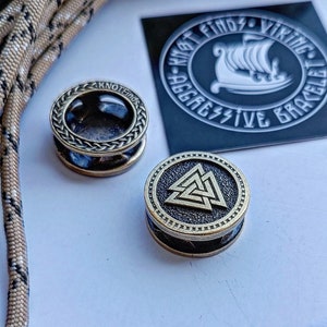 Op de afbeelding: Twee metalen armbandbedels, een met een gevlochten touwontwerp en de tekst "Knot Finds", en de andere met een goudkleurig Valknut-symbool.