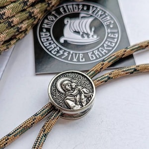 May include: A silver-toned metal bead featuring a detailed image of a religious figure holding a child. The bead is attached to a brown and green paracord bracelet with the text "Knot Finds Viking Aggressive Bracelet" on a black background.