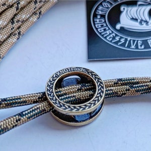 Op de afbeelding: Een close-up van een gouden metalen ring met een gevlochten ontwerp en de woorden "KNOTFinds" gegraveerd aan de zijkant. De ring is geregen met twee strengen camouflage-paracord.