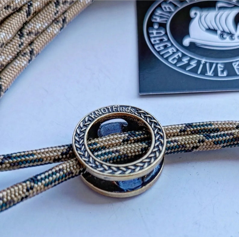 Op de afbeelding: Een close-up van een gouden metalen ring met een gevlochten ontwerp en de woorden "KNOTFinds" gegraveerd aan de zijkant. De ring is geregen met twee strengen camouflage-paracord.