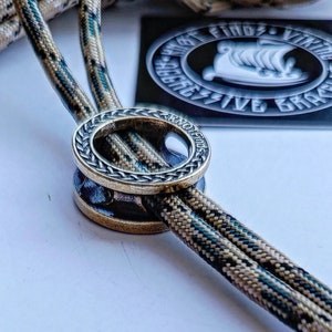 Op de afbeelding: Een close-up van een messing ring met een gevlochten ontwerp en de woorden "Knot Finds" gegraveerd aan de zijkant. De ring is geregen op twee strengen camouflage paracord.