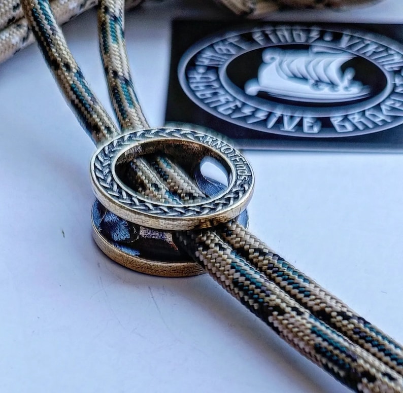 Op de afbeelding: Een close-up van een messing ring met een gevlochten ontwerp en de woorden "Knot Finds" gegraveerd aan de zijkant. De ring is geregen op twee strengen camouflage paracord.