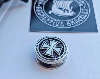 May include: A silver-toned metal bead with a black and white iron cross design. The bead is round and has a textured edge. The bead is part of a bracelet.