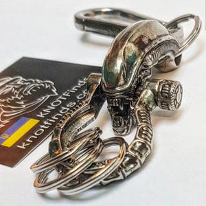 May include: A silver-colored Alien head keychain with intricate details, attached to a key ring and a carabiner clip. The keychain is next to a black card with the text "KNOTFinds.co" and a yellow and blue flag.