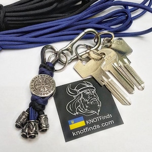 Op de afbeelding: Een sleutelhanger met een zilverkleurige Viking kompas bedel en decoratieve kralen. De sleutelhanger is bevestigd aan een karabijnhaak met sleutels. Blauw en zwart touw is zichtbaar op de achtergrond. Een zwarte kaart met een Viking illustratie en de tekst "KNOTFinds" is ook aanwezig.