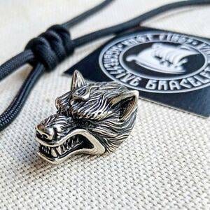 Op de afbeelding: Een zilveren wolfskop-bedel met een gedetailleerd ontwerp. De bedel is bevestigd aan een zwarte paracord-ketting.