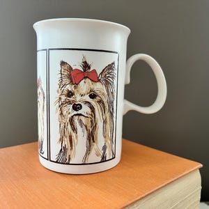 May include: White ceramic mug with a Yorkshire Terrier illustration. The dog has brown and tan fur and a red bow. The mug has a curved handle. The background is a neutral grey. The mug is sitting on a book.
