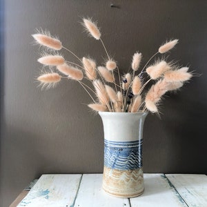 Peut inclure: Un vase en céramique blanche avec un motif ondulé bleu et brun, rempli d'un bouquet de pampas séchées.