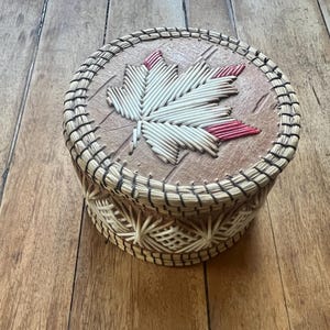 May include: A round, woven basket with a maple leaf design on the lid. The leaf is made of white and red woven material. The basket has a natural, light brown colour with a dark brown woven trim.