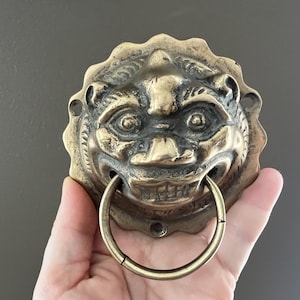 Op de afbeelding: Een messing deurklopper met een gedetailleerd leeuwenkopontwerp en een cirkelvormige handgreep. De klopper heeft een verweerde uitstraling, met ingewikkelde details op het gezicht en de manen van de leeuw. De kleur is een warm, verouderd goud.