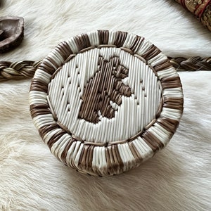 Peut inclure: Un panier rond tissé avec un motif d'ours au centre. Le panier est fait de matériaux de couleur naturelle, avec une silhouette d'ours brun sur un fond blanc. Le bord et le bord extérieur sont bruns. Le panier est posé sur une surface blanche ressemblant à de la fourrure.