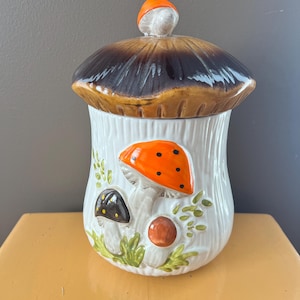 Può includere: Un barattolo in ceramica a forma di fungo con coperchio. Il barattolo è bianco con funghi arancioni e marroni dipinti e foglie verdi. Il coperchio è marrone e beige con un piccolo fungo arancione sopra.