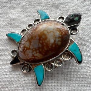 May include: A silver-toned turtle brooch with a brown and white shell, turquoise flippers, and black head. The brooch has a detailed design with small, round silver accents around the shell and flippers. A unique piece of jewelry.
