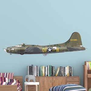 Puede incluir: Un adhesivo de pared de un avión B-17 Flying Fortress verde y marrón en la habitación de un niño. El adhesivo está en una pared azul claro sobre una cama de madera con ropa de cama a rayas. Hay estanterías y un escritorio con una silla en la habitación.