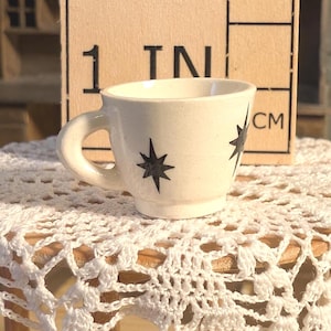 Puede incluir: Una pequeña taza de té de cerámica blanca con diseños de estrellas negras, colocada sobre una mesa de madera en miniatura cubierta con un mantel individual de encaje blanco. La mesa mide 2,54 cm de altura, como indica el texto en un bloque de madera detrás de la taza.