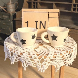 Puede incluir: Dos pequeñas tazas de cerámica blanca con diseños de estrellas negras sobre una pequeña mesa redonda cubierta con un mantel individual de encaje blanco. La mesa es de madera. Una caja de cartón con el texto "1 IN" está detrás de las tazas.