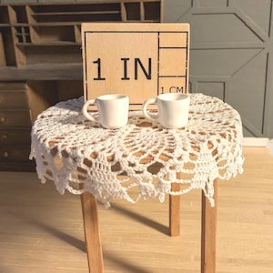 Pode incluir: Duas pequenas canecas de cerâmica branca estão sobre uma mesa de madeira em miniatura coberta por uma toalha de crochê branca. Um bloco de madeira atrás das canecas diz "1 IN" e "1 CM". A mesa tem quatro pernas de madeira.
