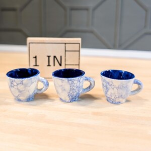Puede incluir: Tres pequeñas tazas de café de cerámica azul y blanca con textura burbujeante. Las tazas están alineadas en una superficie de madera con una regla que dice "1 IN" en el fondo.