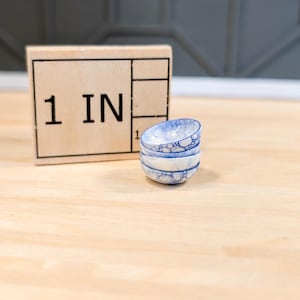 Puede incluir: Un conjunto de cuatro cuencos pequeños de cerámica azul y blanca con un esmalte burbujeante. Los cuencos están apilados sobre una superficie de madera con una regla que dice "1 IN" en el fondo.