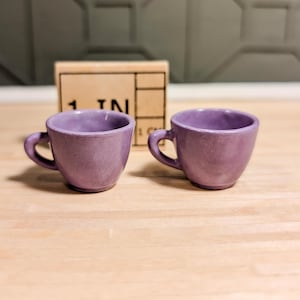 Puede incluir: Dos pequeñas tazas de té de cerámica de color púrpura con asas redondeadas. Las tazas están sobre una superficie de madera marrón claro.