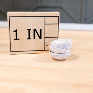 Puede incluir: Un letrero de madera con el texto "1 IN" en él, junto a una pila de cuatro cuencos pequeños de cerámica blanca con un patrón moteado.