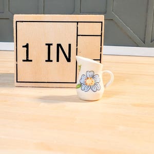 Puede incluir: Una pequeña jarra de cerámica blanca con un diseño floral azul y amarillo. La jarra mide 1 pulgada de alto.