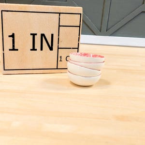Puede incluir: Un conjunto de cuatro cuencos pequeños de cerámica blanca con un borde rosa. Los cuencos están apilados sobre una superficie de madera. El texto "1 IN" es visible en un bloque de madera en el fondo.