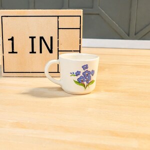 May include: A white ceramic teacup with a blue floral design. The cup has a handle and is 1 inch tall.