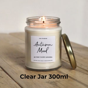 May include: A clear glass jar candle with a gold lid. The candle is labeled "Autumn Mood" and "Buttery Pastry & Raisins". The candle is lit and the flame is visible. The jar is 300 ml.