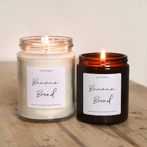 May include: Two lit glass jar candles with the text "Banana Bread" on white labels. One candle is in a clear jar, the other in a brown jar. The text "Natural Wax Candle Made in England" is below the main text.