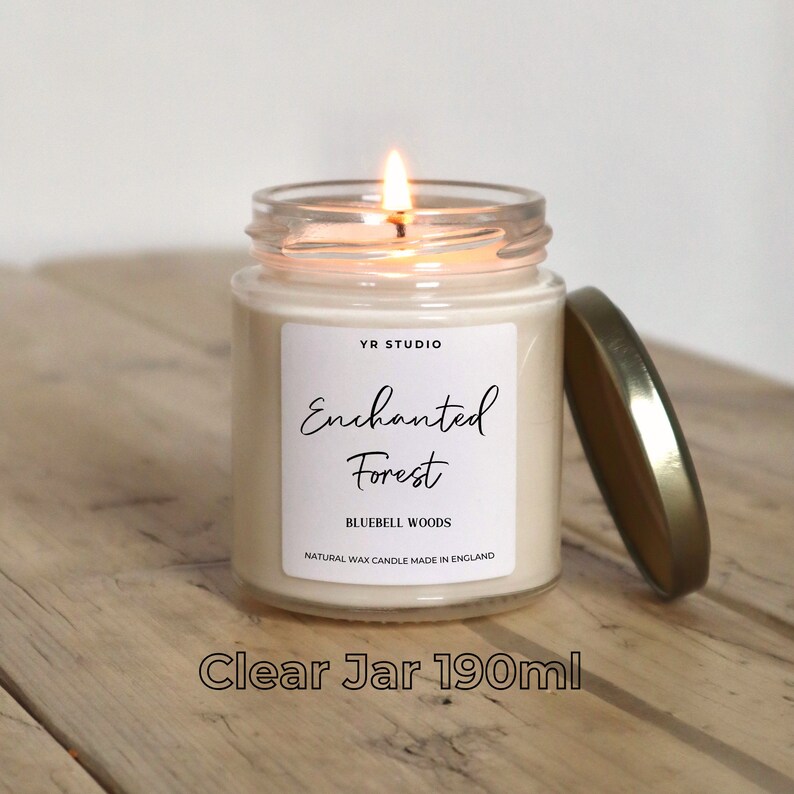 Puede incluir: Una vela en un tarro de vidrio transparente con tapa dorada. La vela est&aacute; etiquetada con "YR STUDIO Enchanted Forest Bluebell Woods Natural Wax Candle Made in England". La vela est&aacute; encendida y la llama es visible. El tarro es de 190 ml.