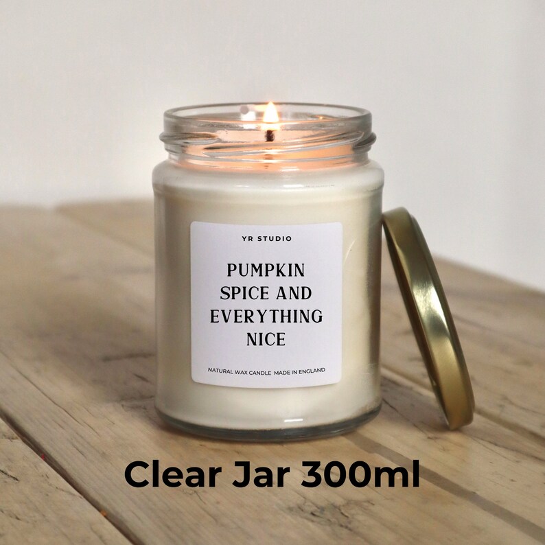May include: A clear glass jar candle with a white label that reads "YR STUDIO Pumpkin Spice and Everything Nice Natural Wax Candle Made in England". The candle is lit and the jar is sitting on a wooden surface. The candle is 300 ml.