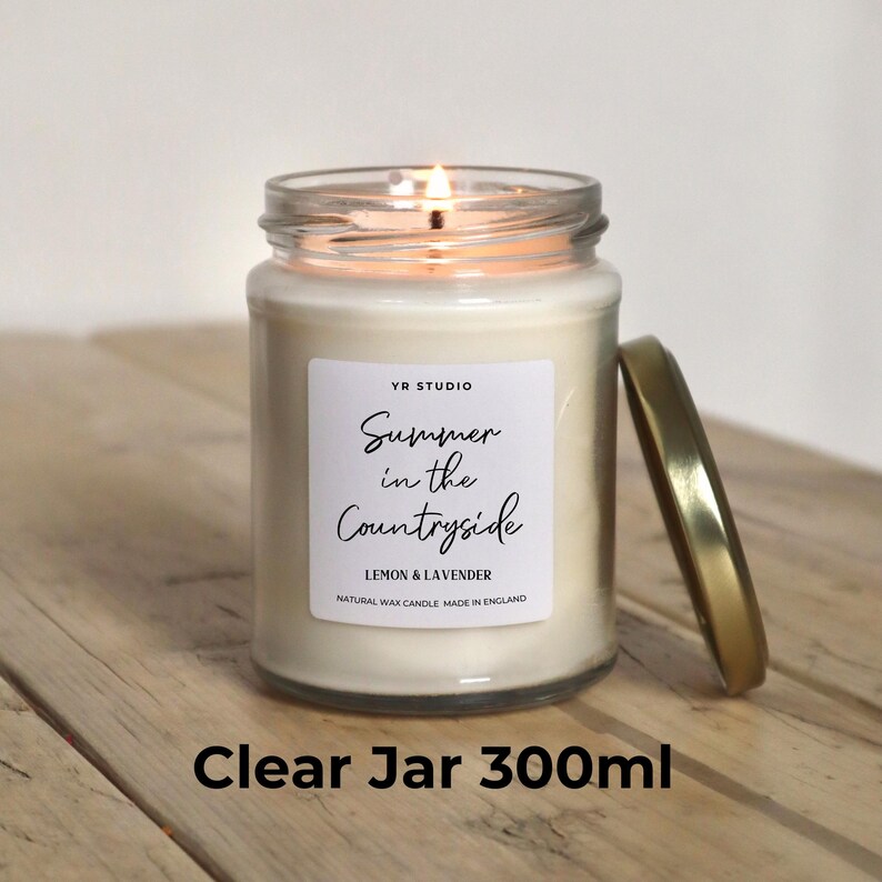 May include: A clear glass jar candle with a white label that reads "YR STUDIO Summer in the Countryside Lemon & Lavender Natural Wax Candle Made in England". The candle is lit and the jar is sitting on a wooden surface. The jar is 300 ml.