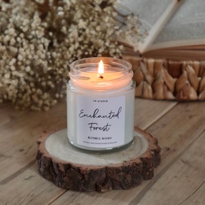 Puede incluir: Una vela blanca en un tarro de vidrio con una tapa de madera. La vela est&aacute; encendida y la llama es visible. El tarro tiene una etiqueta blanca con el texto "YR Studio Enchanted Forest Bluebell Woods Natural Soy Candle Made in England".