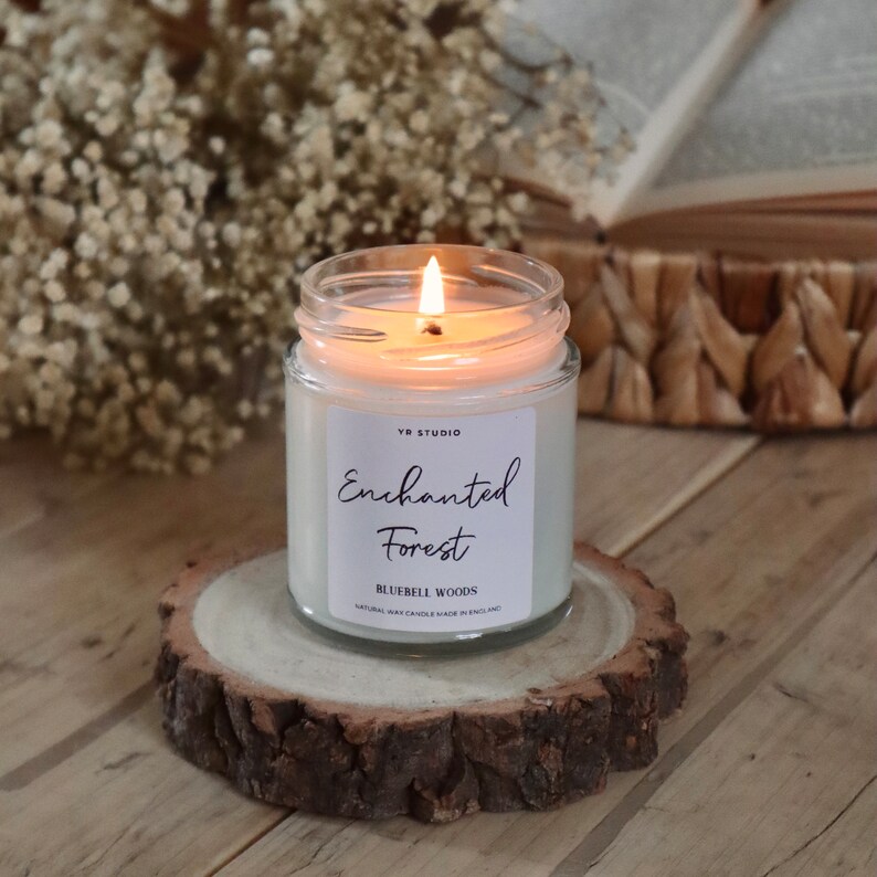 Puede incluir: Una vela blanca en un tarro de vidrio con una tapa de madera. La vela est&aacute; encendida y la llama es visible. El tarro tiene una etiqueta blanca con el texto "YR Studio Enchanted Forest Bluebell Woods Natural Soy Candle Made in England".