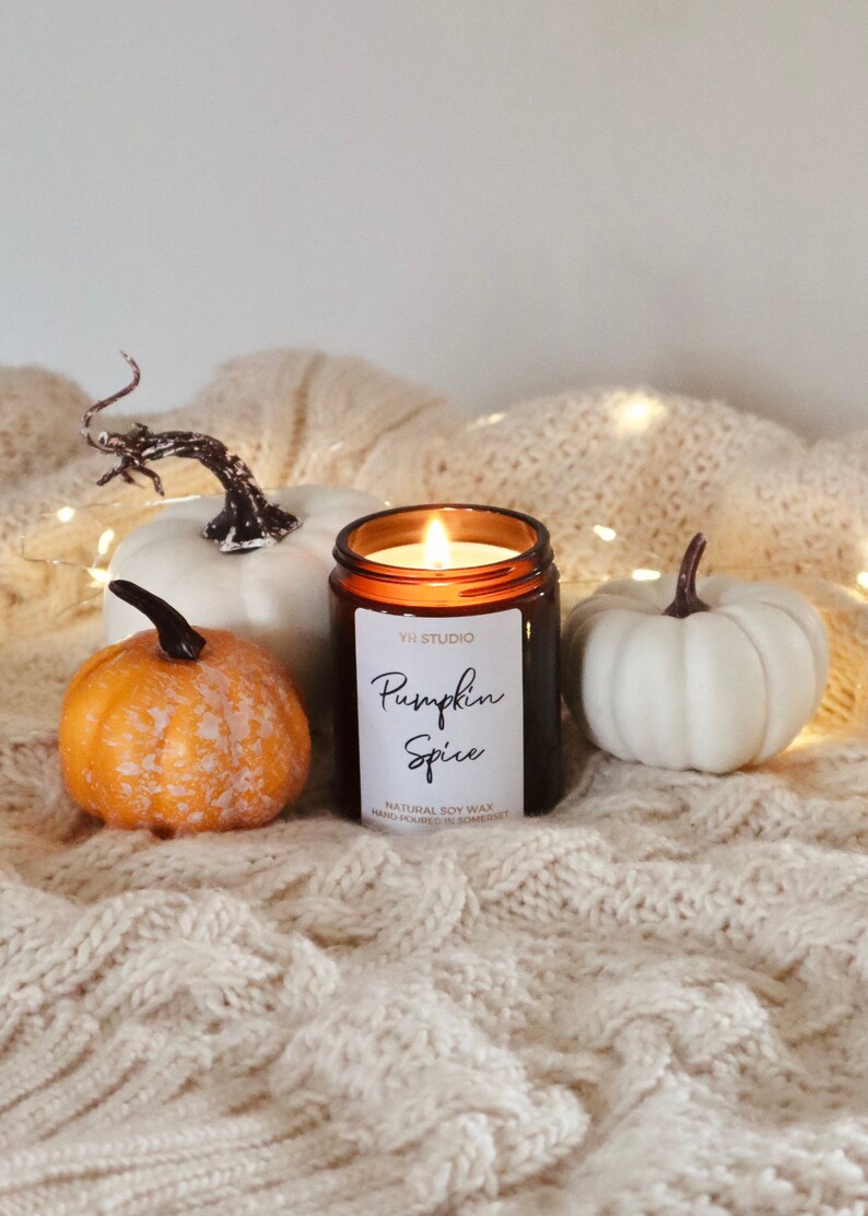 May include: A brown glass candle with a white label that reads "Yi Studio Pumpkin Spice Natural Soy Wax Hand Poured in Somerset" sits on a white knitted blanket with two white pumpkins and one orange pumpkin.