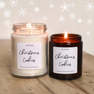 May include: Two lit glass jar candles with the text "Christmas Cookies" on white labels. One candle is in a clear jar with a white wax, and the other is in a brown jar with a brown wax. The background has snowflake decorations.