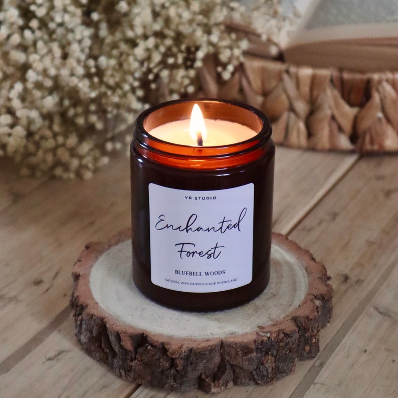 Puede incluir: Un tarro de vela de vidrio marr&oacute;n con una etiqueta blanca que dice "Enchanted Forest" y "Bluebell Woods" con una vela de cera natural hecha en Inglaterra. La vela est&aacute; encendida y la llama es visible.