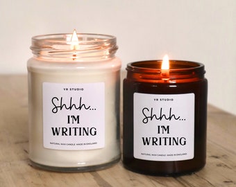 May include: Two candles with brown glass jars and white labels. The labels have the text "Shhh... I'M WRITING" and "NATURAL WAX CANDLE MADE IN ENGLAND".