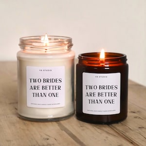 May include: Two glass candle jars with brown lids. Both candles have white labels with the text "Two Brides Are Better Than One" and "YR Studio" printed on them. The candles are lit and the flames are visible.